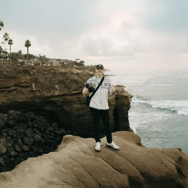 Chase standing on a coastal cliff with his camera, ocean behind him.